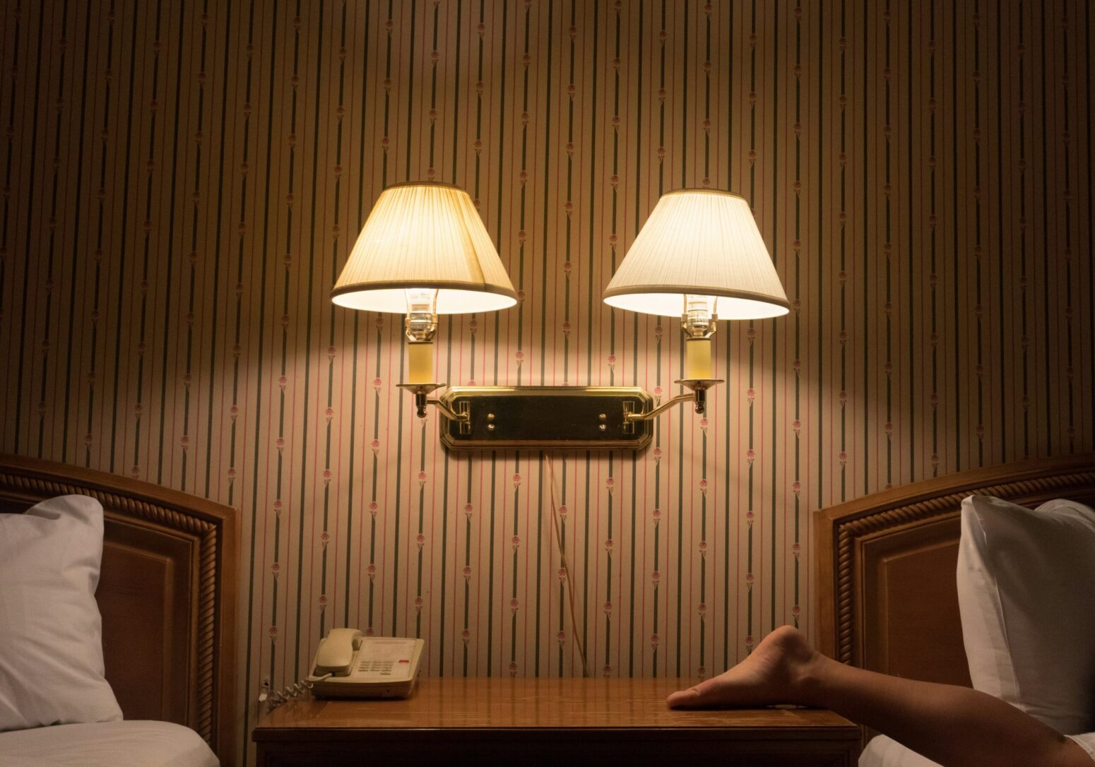 Two lamps mounted on the wall of a motel room. There are beds on either side of the image, a phone on the table below the lamps, and a person's foot on the right side of the table.