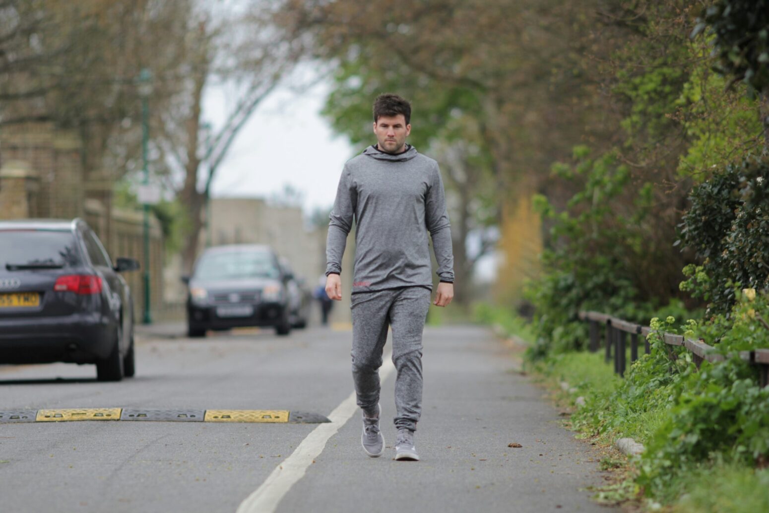 A white man in long-sleeved gray shirt and sweatpants walking on the side of the road with cars blurred to the left and greenery to the right