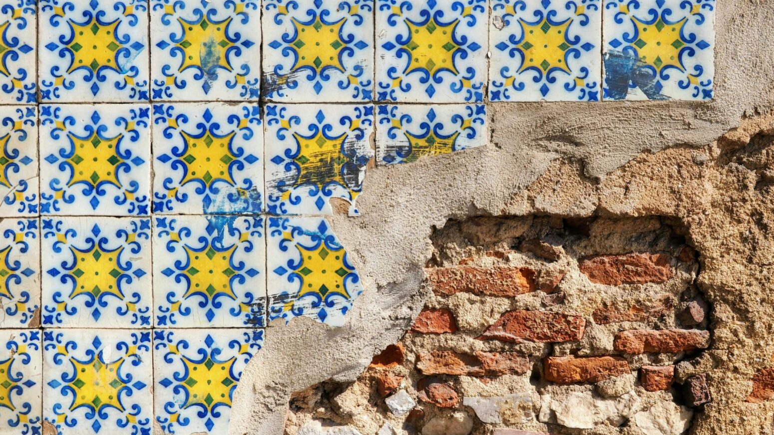 A wall of blue and yellow tiles, some of which has been torn away and reveals older brick and stone