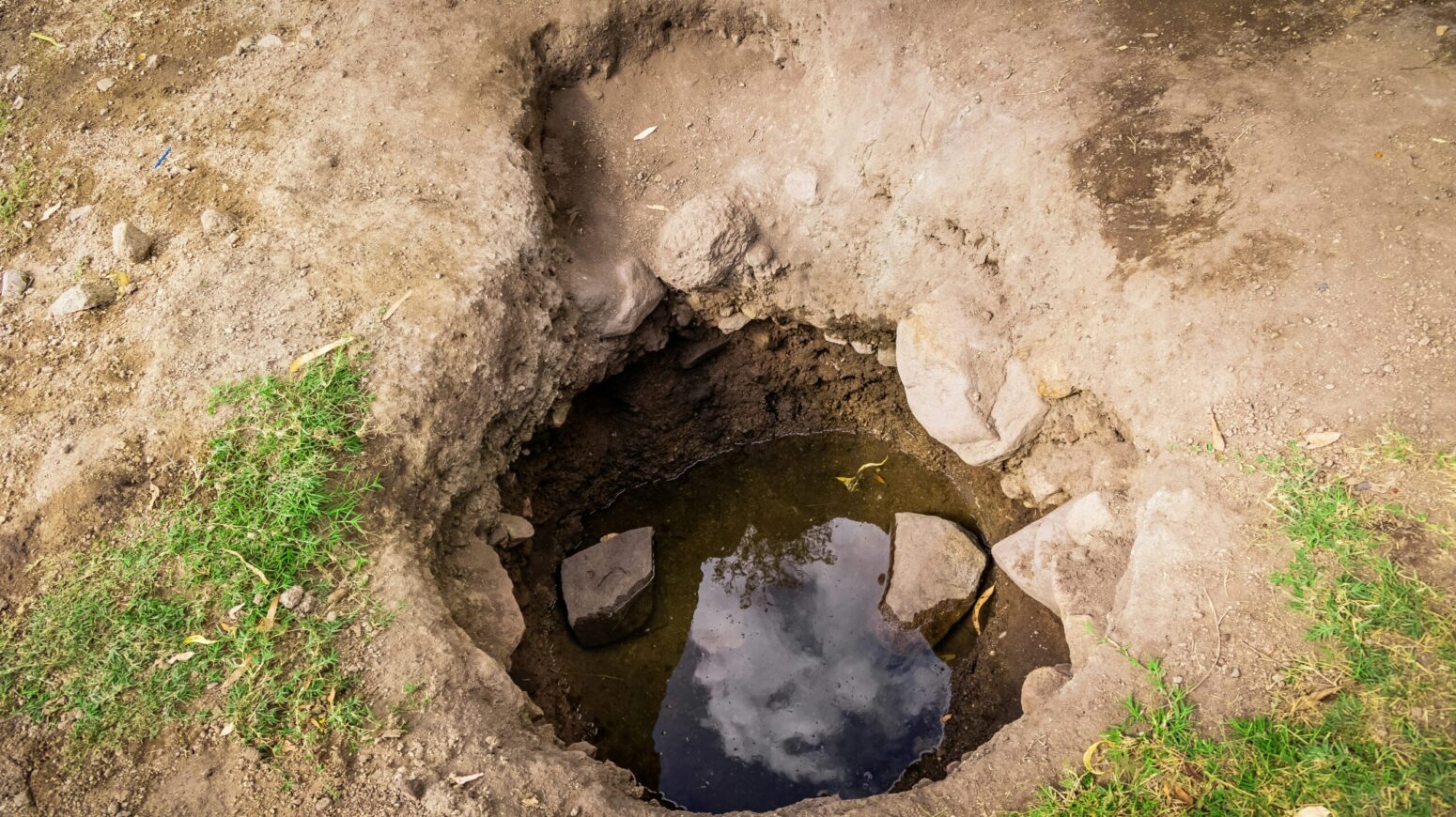 A hole dug into the ground with a small amount of grass on either side and the sky reflected in water within the hole