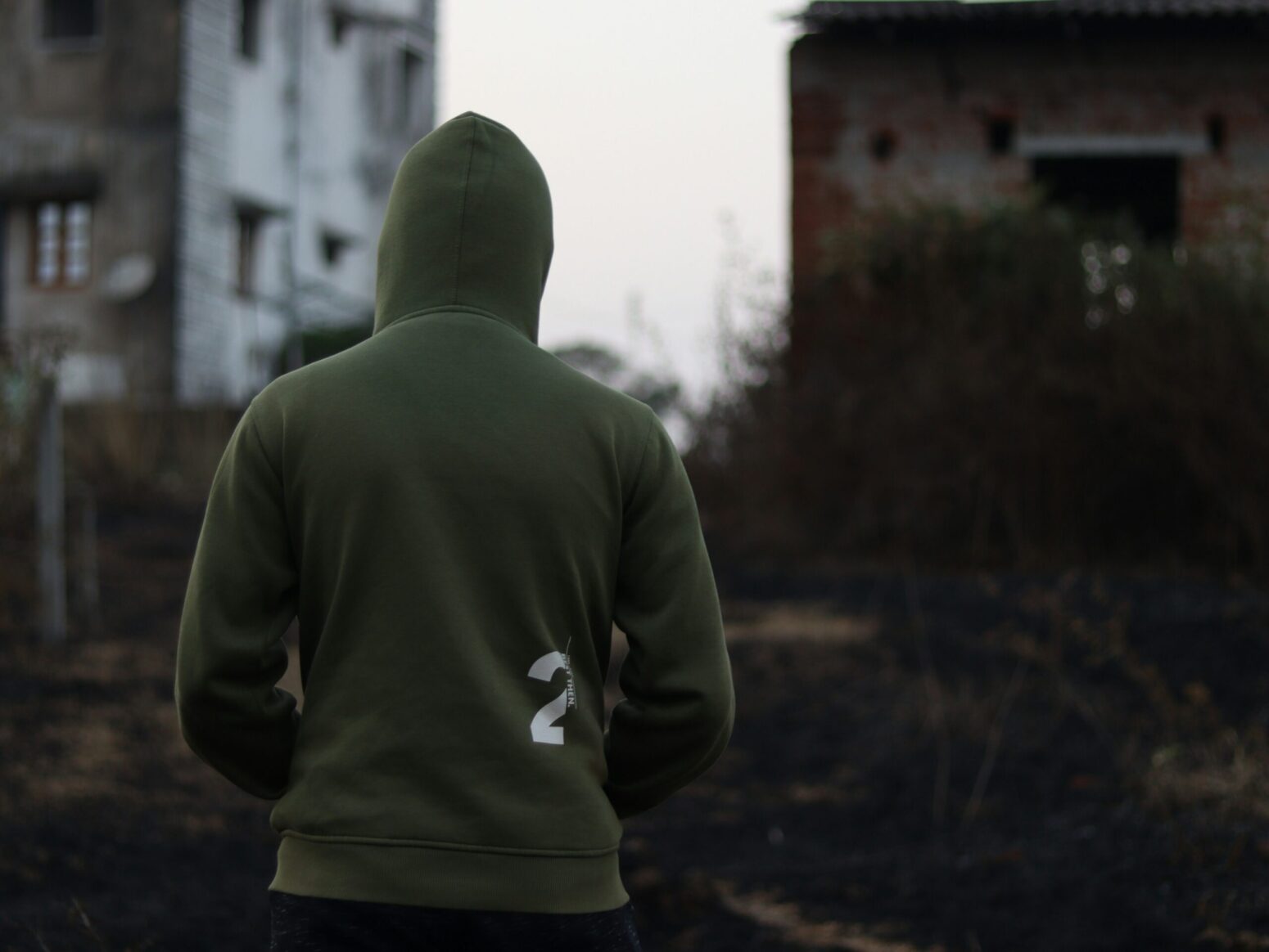 The back of a person in a green hoodie with the hood up and their hands in their pockets. Buildings are blurred in the background