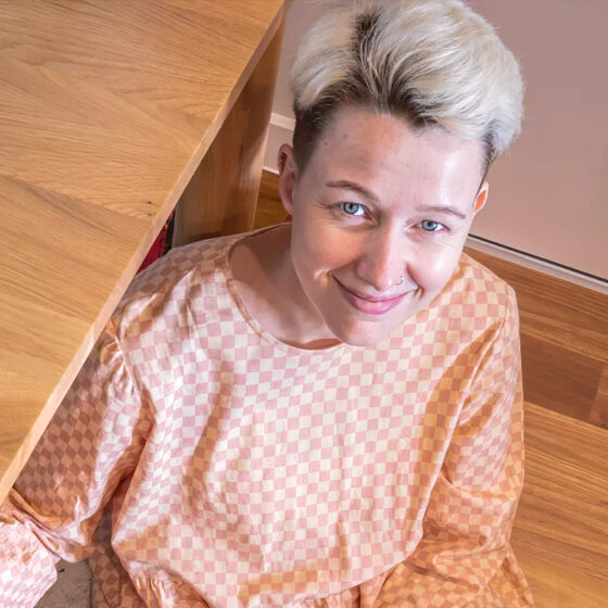 Eleanor, a white person with short bleached hair wearing a peach-and-white top, looks up at the camera and smiles.