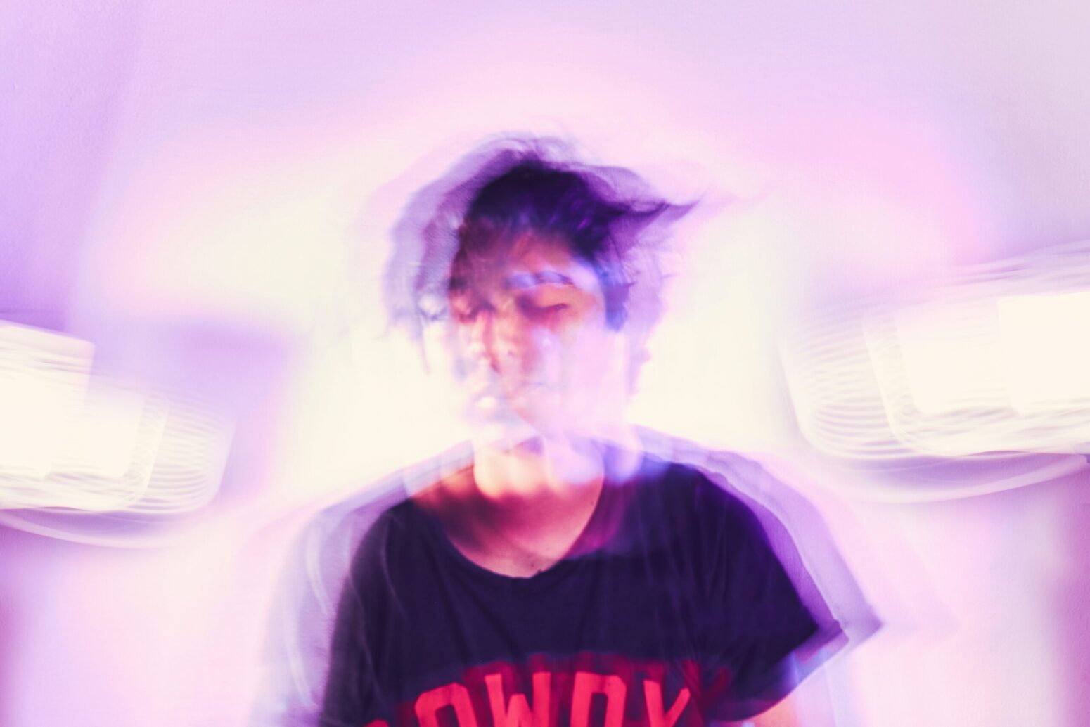 This blurred photograph shows a man in the middle wearing a black shirt with red text on it. The background is a blend of purple and pink.
