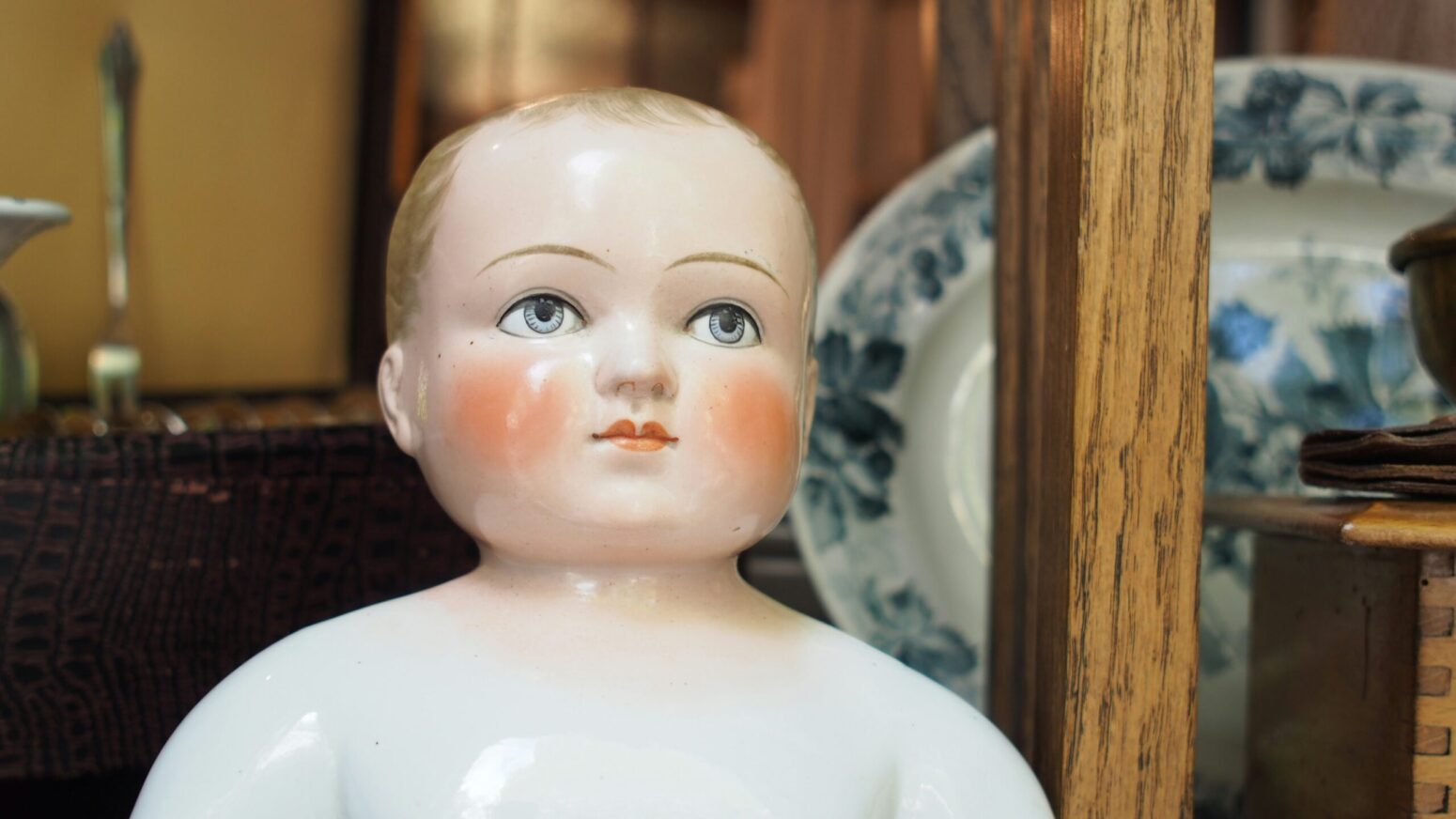 This photograph shows a baby ceramic figurine on a white ceramic plate. This photograph also catches a blue plate, wooden pole, a candle, among other objects in the background. baby ceramic figuring on white ceramic plate