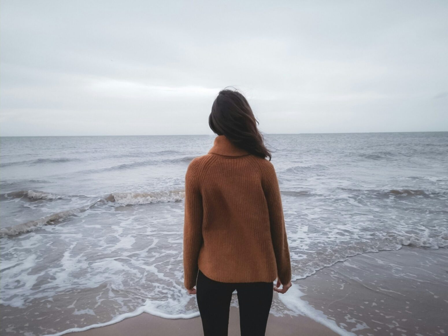 This photograph shows a woman looking out into the ocean on a cloudy day.