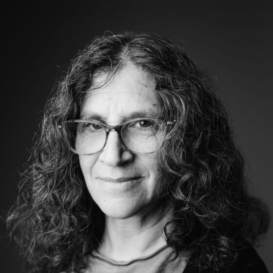 D. Dina Friedman, a white woman with curly hair and square glasses, poses in a moody black-and-white photo.