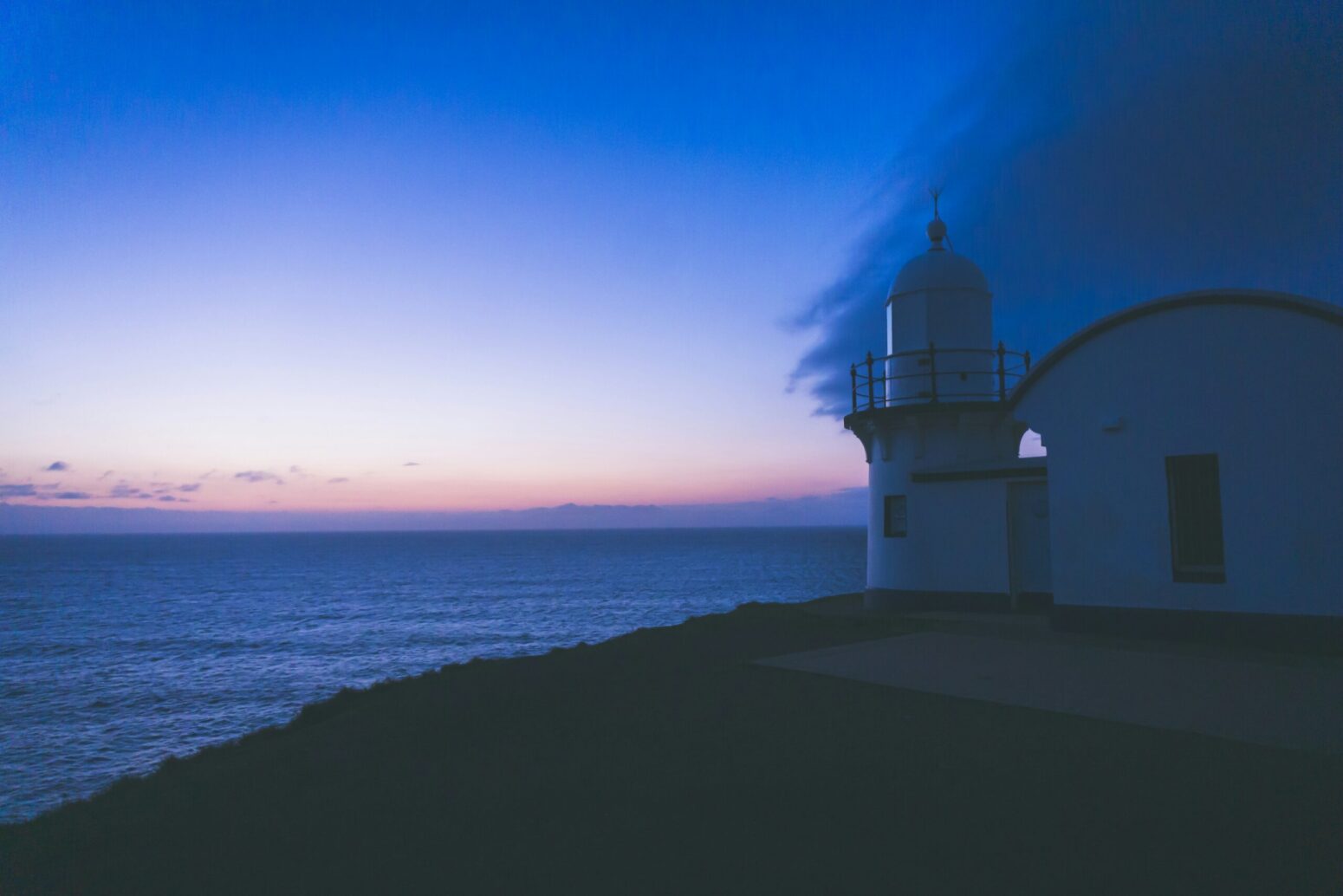 This photograph shows a white building with the ocean in the background. The sunrise illuminates the nightlight ready to succumb over the ocean and land.