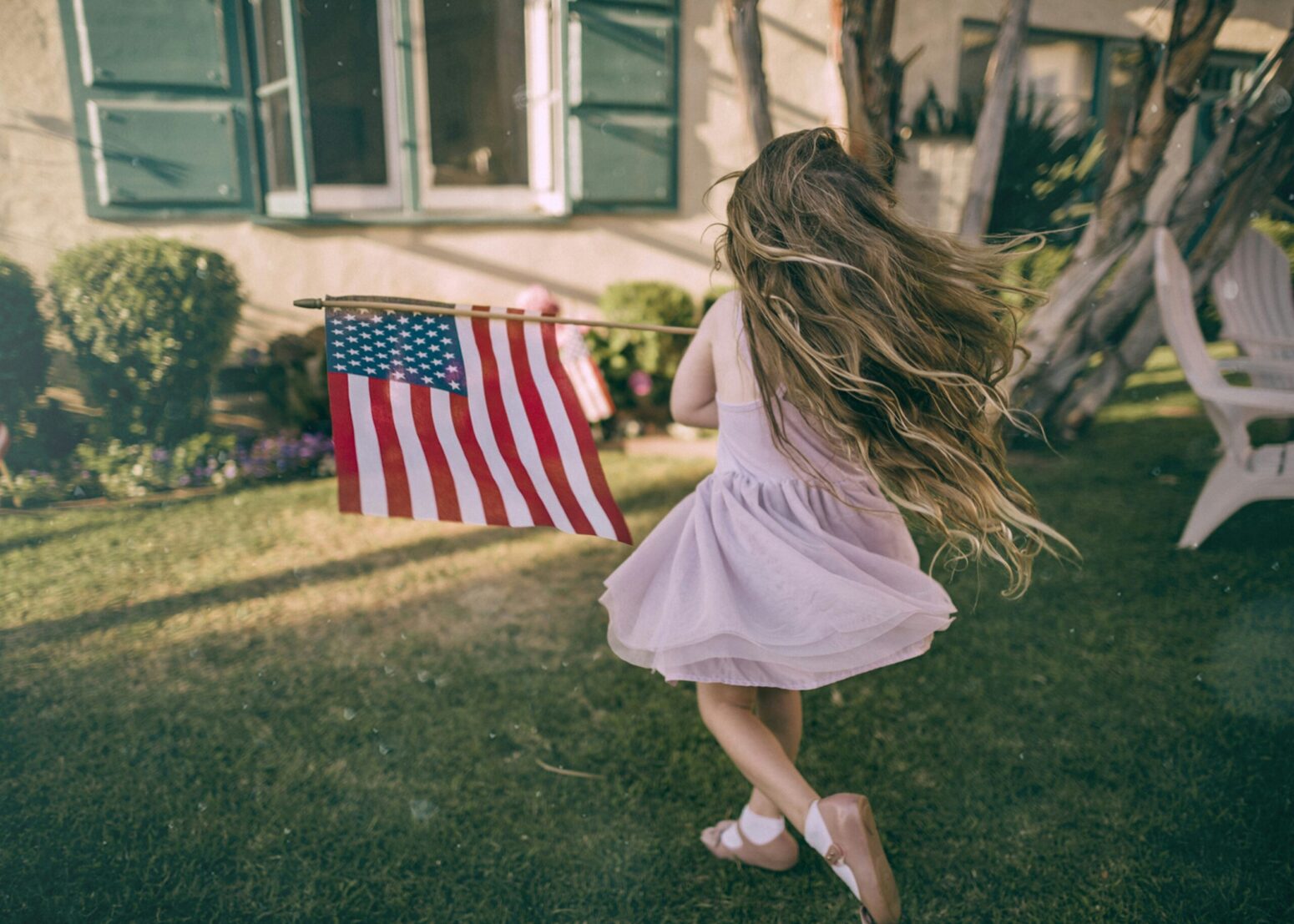 This photograph shows a girl holding an American flag in front of a house, in the front yard. There is sunlight shining on the window and panes.