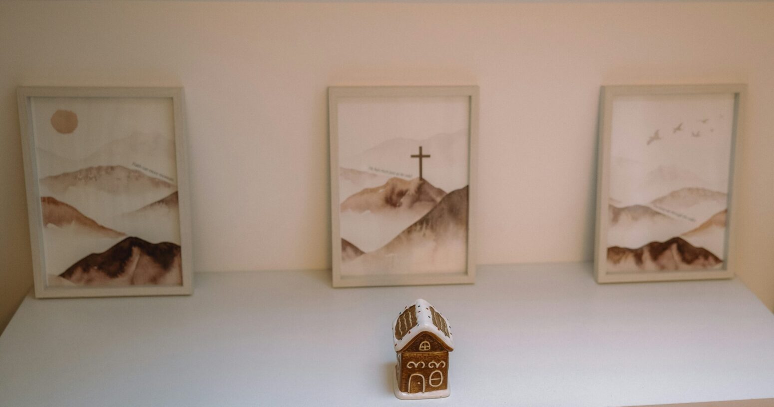 This photograph shows a white counter, with a gingerbread house in the middle, and three pictures evenly spread apart, like a triptych of a hill with a cross in the middle.