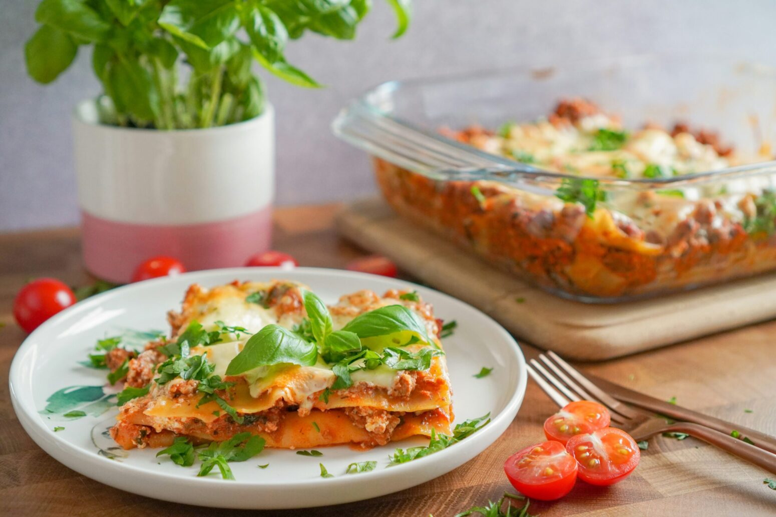 This photograph shows a table with a tray of lasagna, and a fork, tomatoes, and a plant.