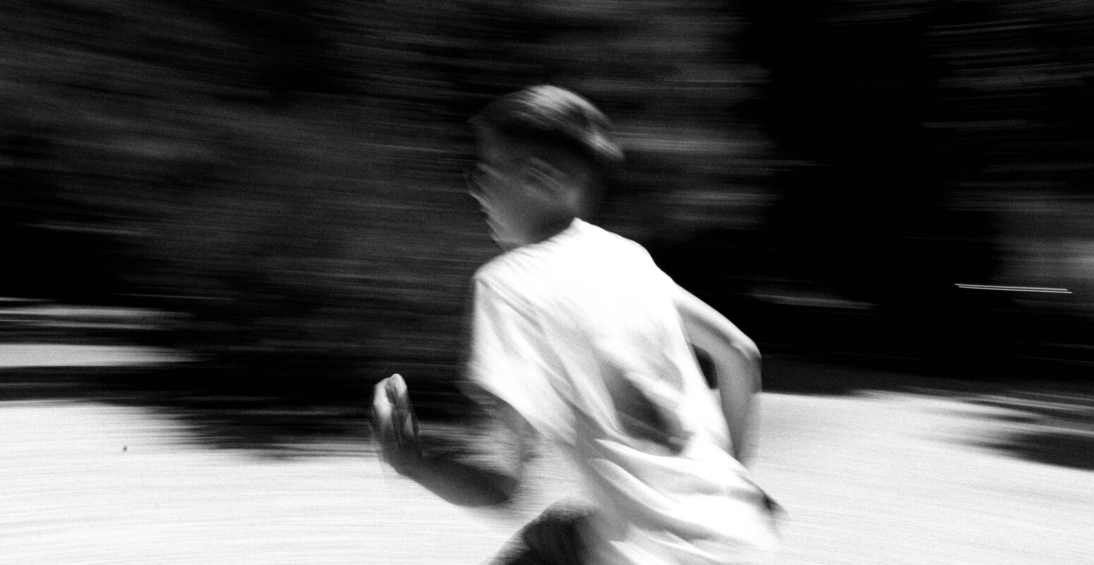 A blurry black and white photo of a boy running in the street. There appears to be trees and signs of nature behind him. The boy is wearing glasses.