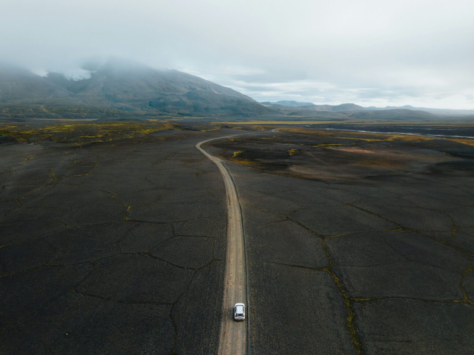 This photograph is an aerial view of a white vehicle driving down a road. There are mountains in the background, visible fog, clouds, and spots of water in the photograph.