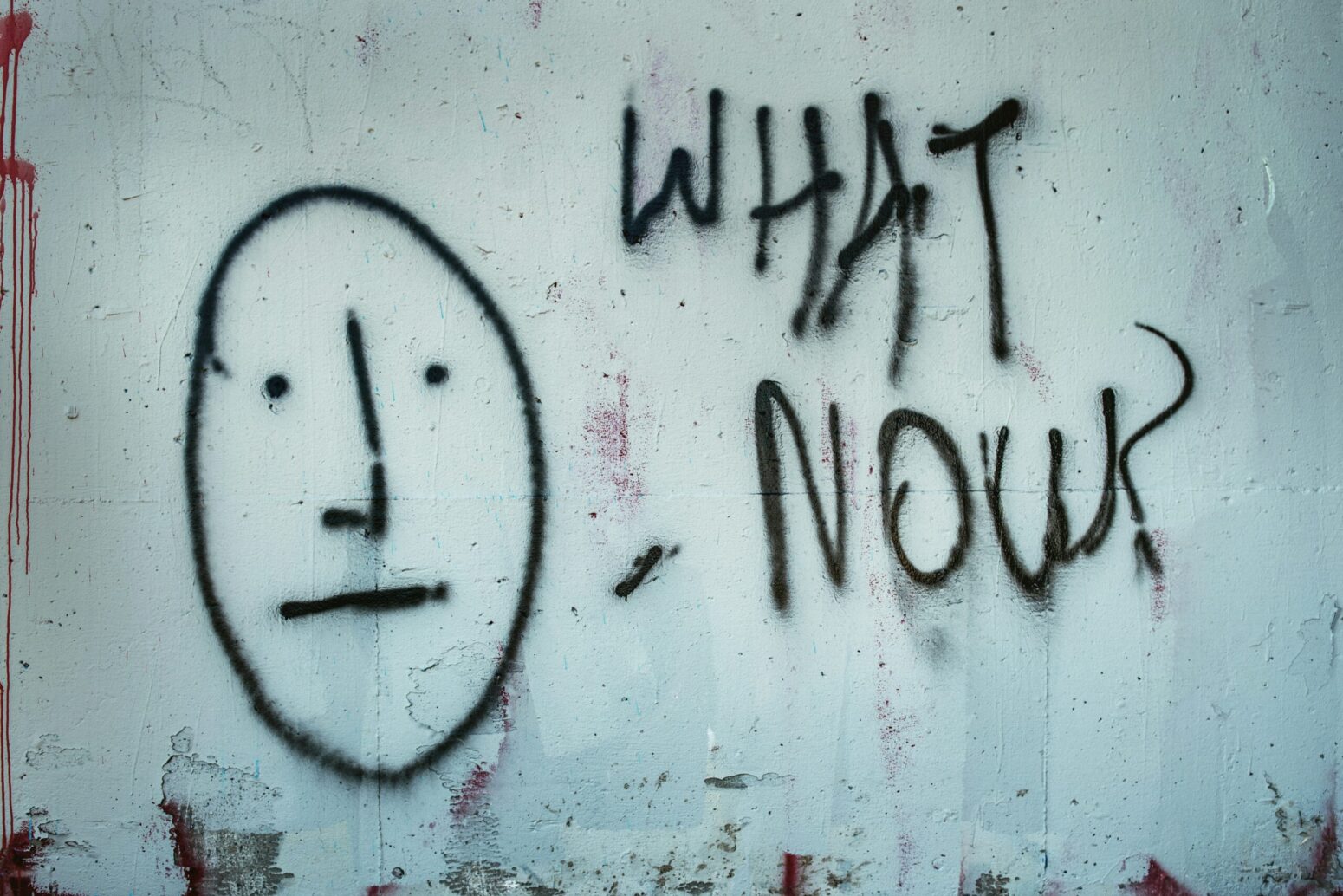 This photograph shows graffiti on a wall, with a black careless face with the words "what now?"