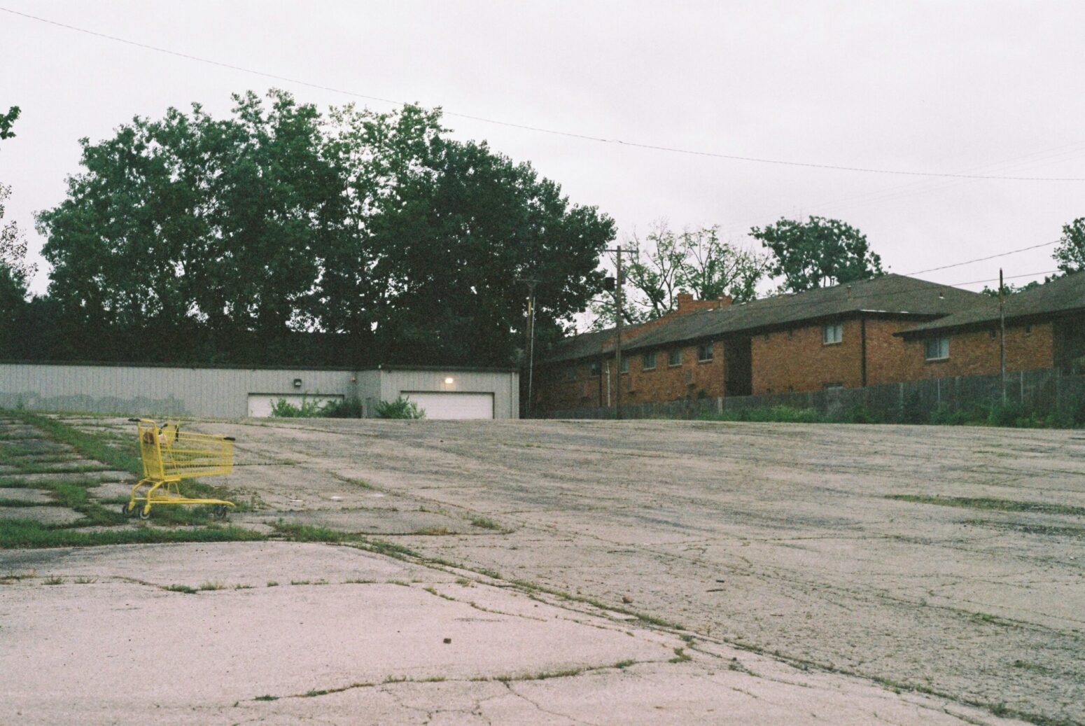 This photograph shows an empty and desolate parking lot with a yellow cart and buildings in the background.