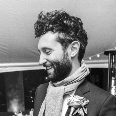 Eric Weiskott, a white man with dark curly beard and hair wearing a suit jacket, corsage, and scarf, smiles in profile. The photo is black-and-white.