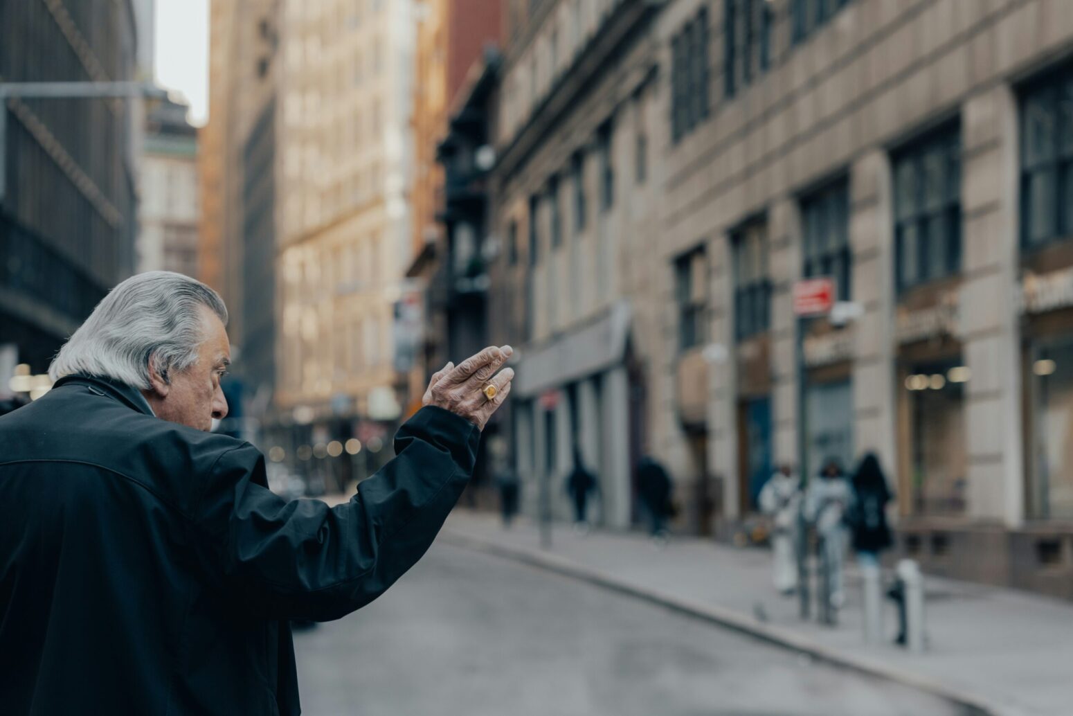 This photograph shows a city in the background with an elderly man waving to another individual not seen within the photograph.