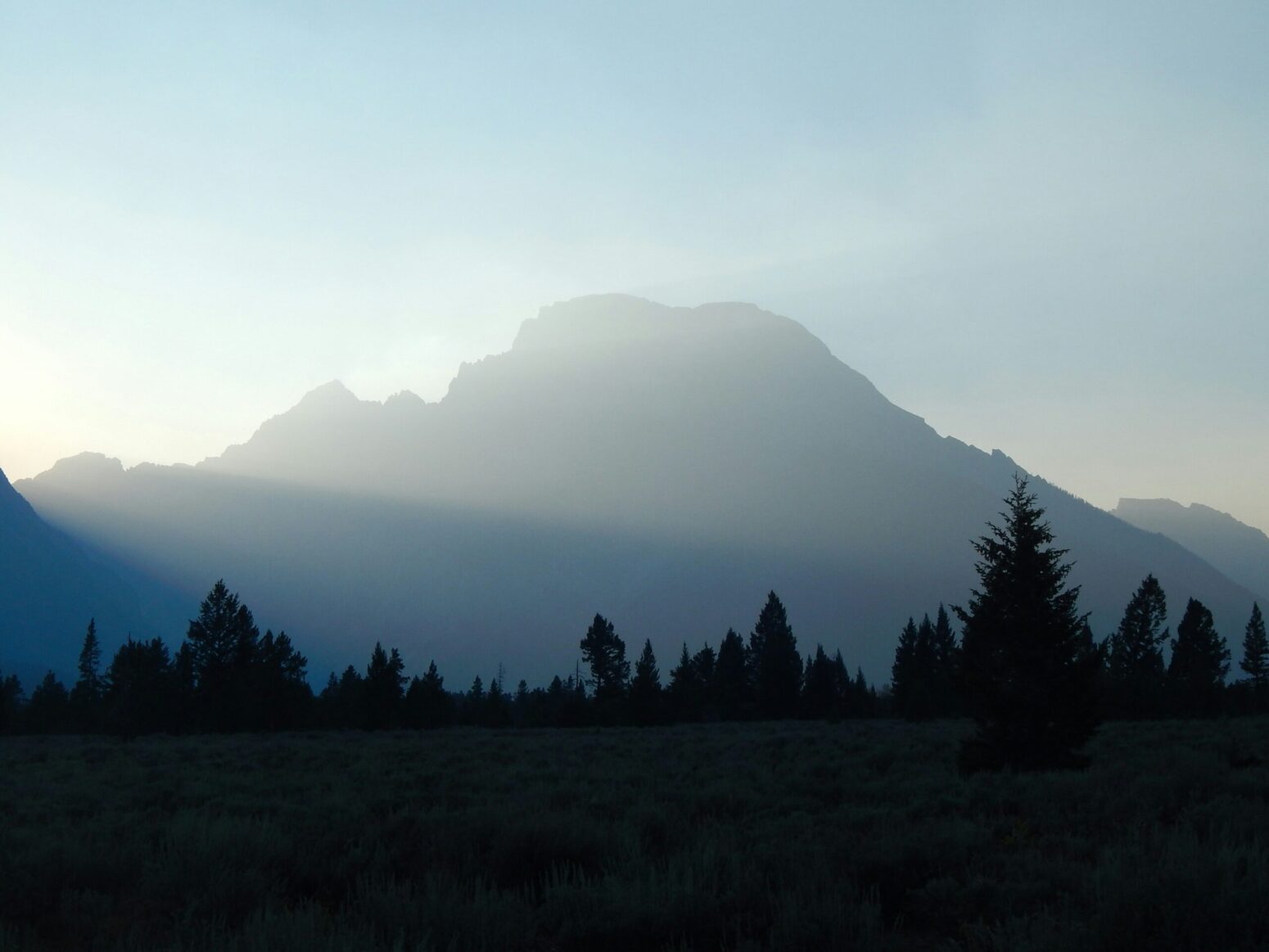 This photograph shows the silhouette of the forest with the sun shining on the mountains within the background.