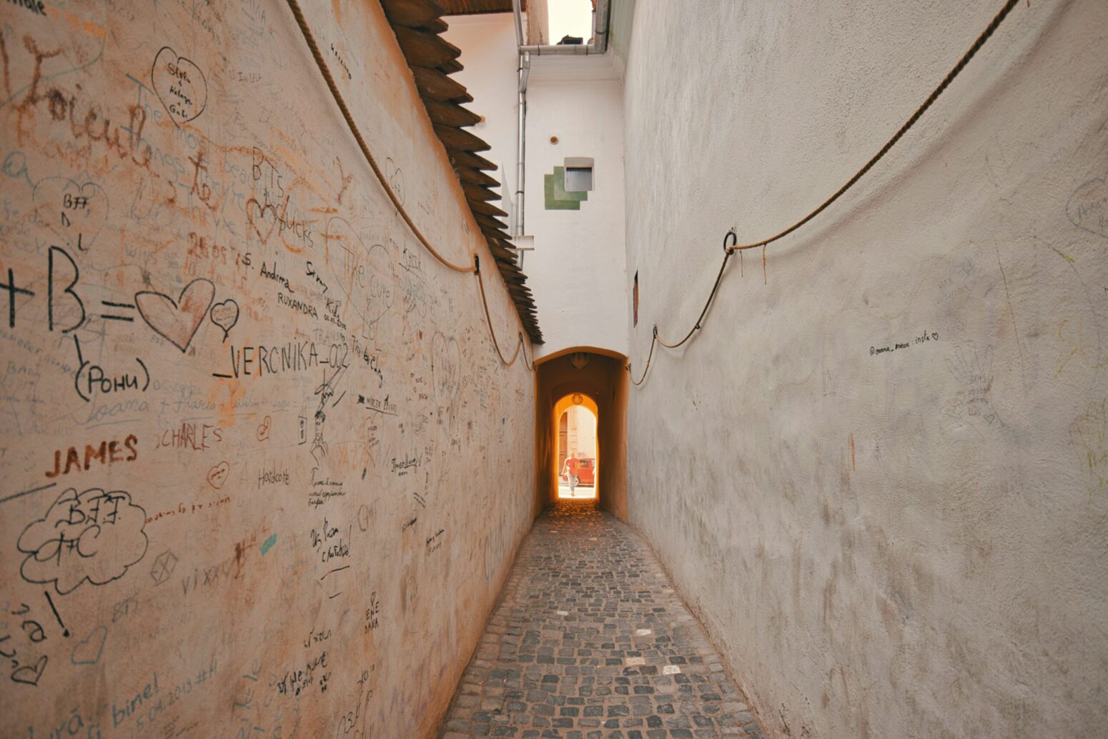This photograph shows a graffitied beige wall and a narrow street with something golden glowing at the end of the narrow street.