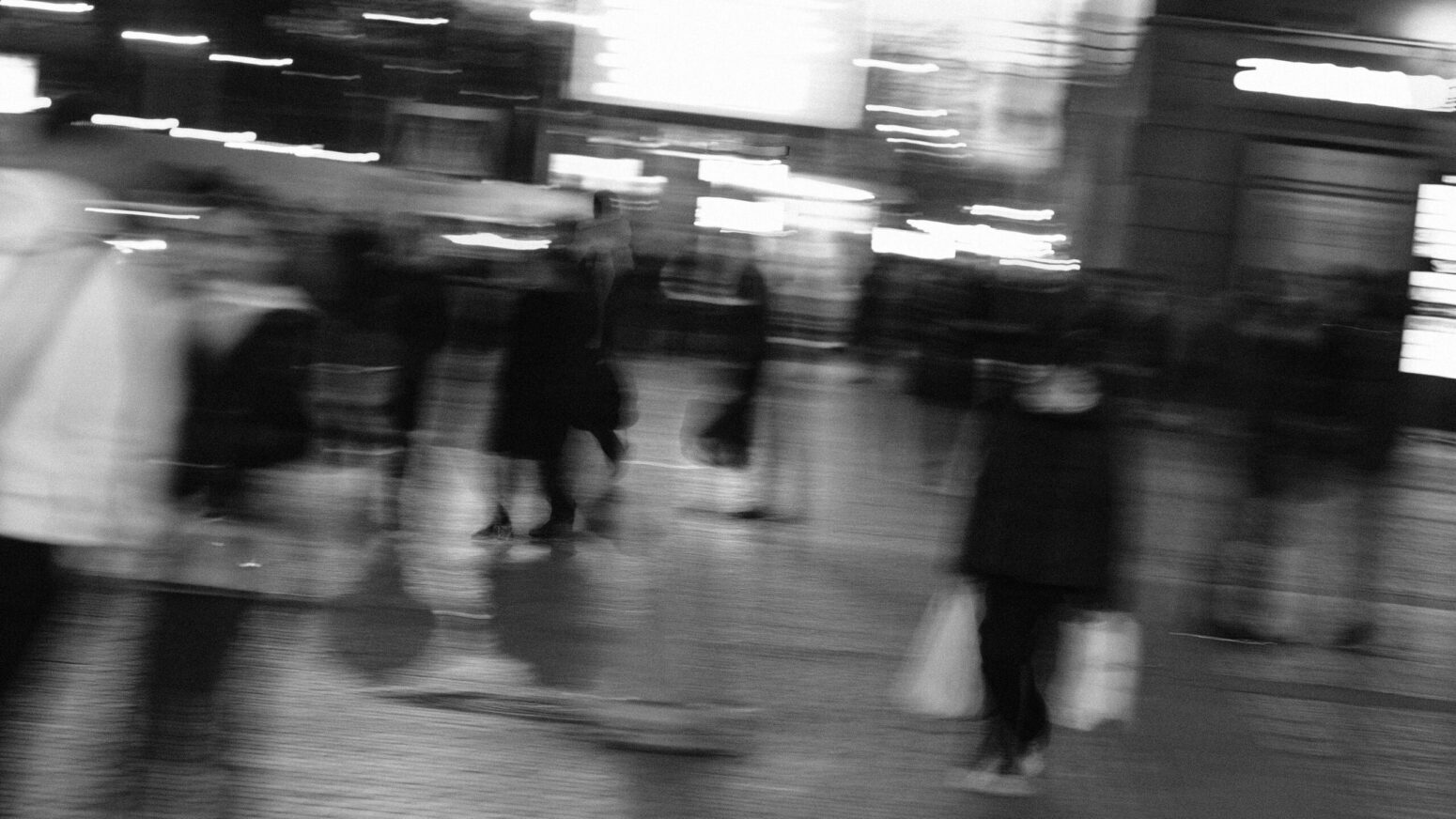 This blurry black and white photograph shows many people within the picture, with buildings, lights, and windows in the background.