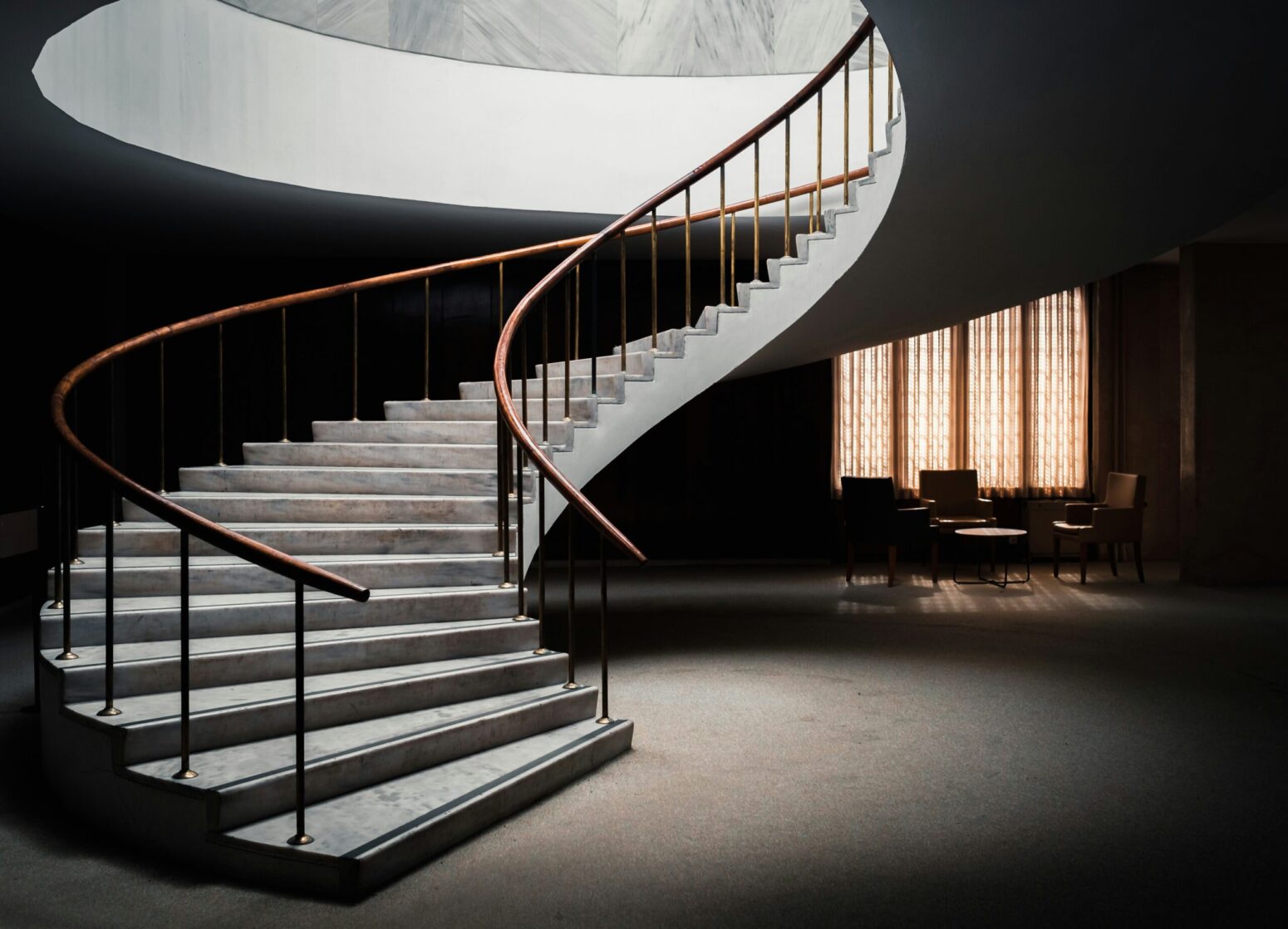 This photograph shows a spiral staircase and a dark room, presumably in the basement, with a closed yellow curtain and a table with a few chairs.