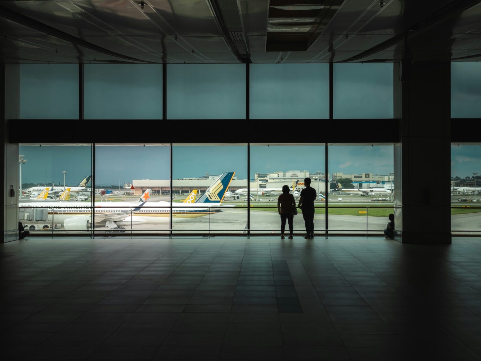 This photograph shows two individuals looking out of the window of an airport, where an airplance can be seen.
