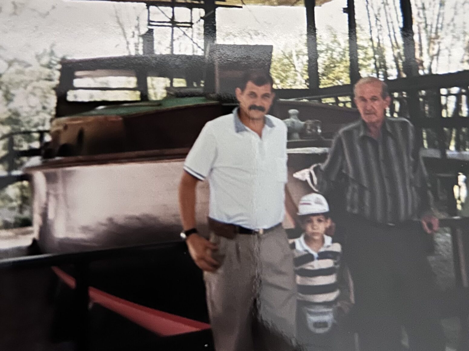 Three Cuban people standing in front of a mahogany brown boat. The man on the left is in his thirties or forties and has a mustache. The one in the middle is a child with a striped shirt and a ballcap. The one on the right is a man older than sixty with a long-sleeve dark striped shirt. The image has a slight glossiness to it