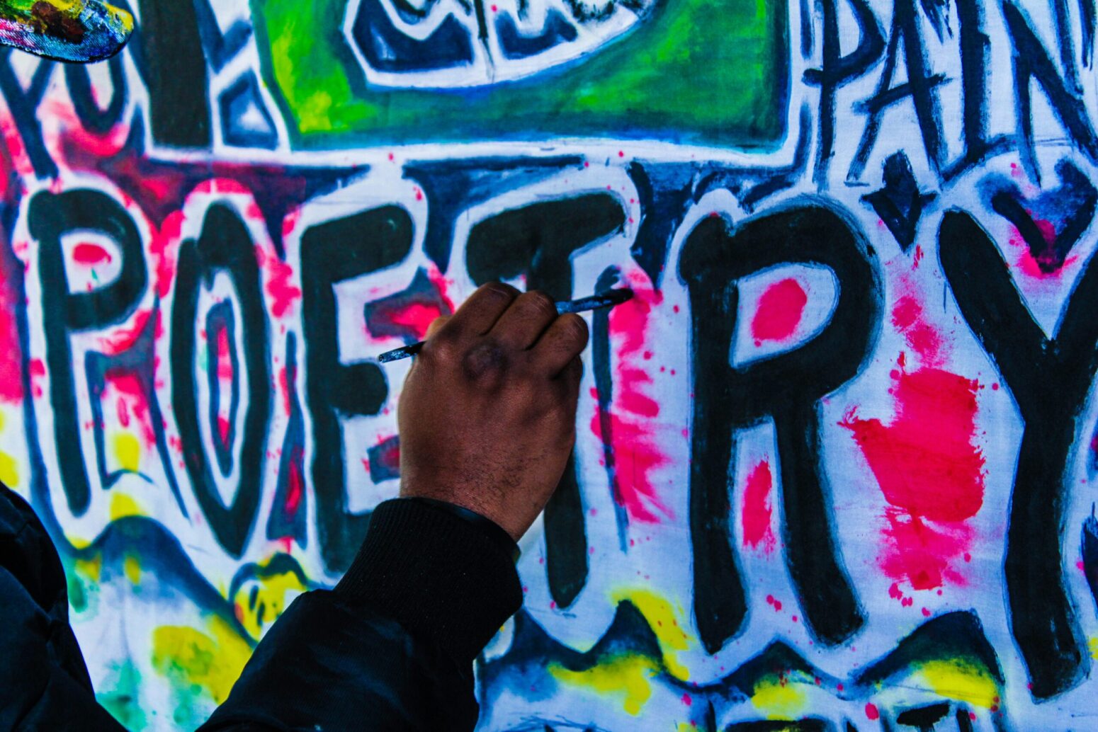 This photograph shows a graffitied wall with the word "POETRY" being scrabbled on the wall, with a hand reaching out towards the letter T.