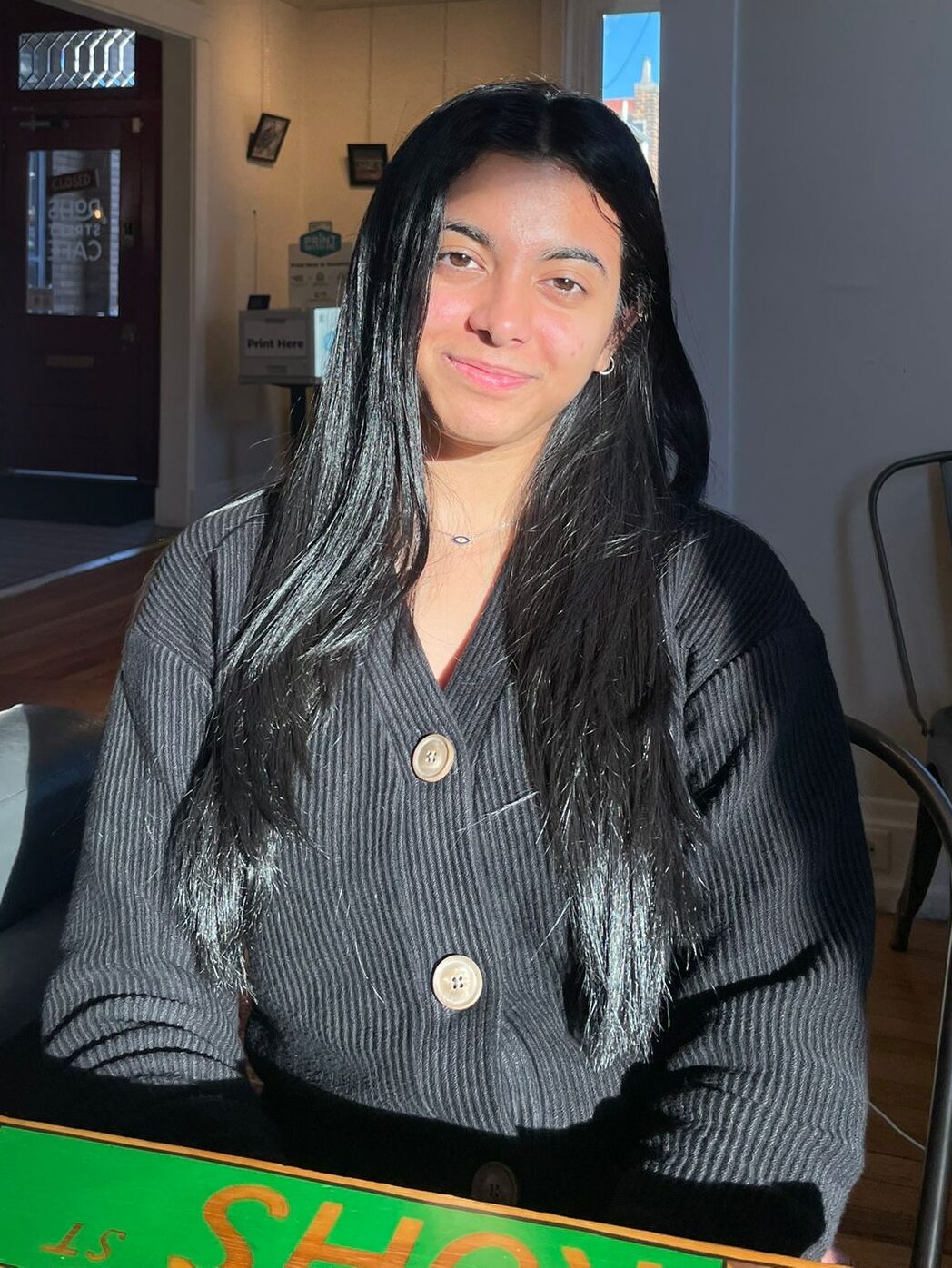Sharvaree Bhalerao, a young Indian woman with long black hair, wears a black cardigan and sits at a table. Sun is shining on her face.