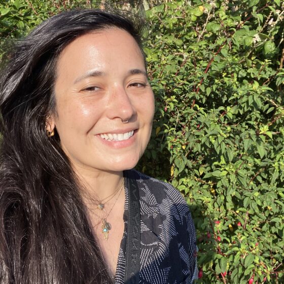 Alison Lubar, an Asian American person with long dark hair, smiles in front of greenery.