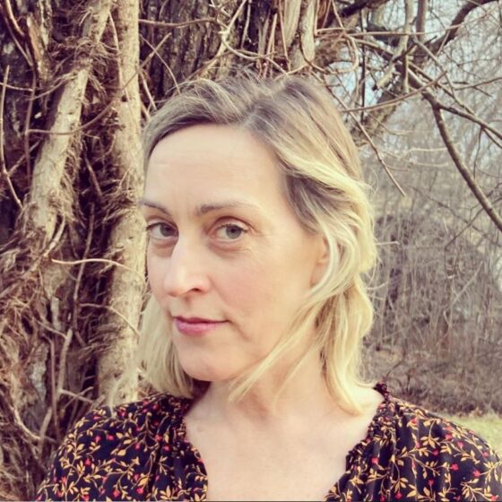 Cheryl Pappas, a white woman with blond hair pulled into a ponytail with a fringe, stands outside against a bare tree wearing a black-and-red floral top. She glances at the camera from the side.