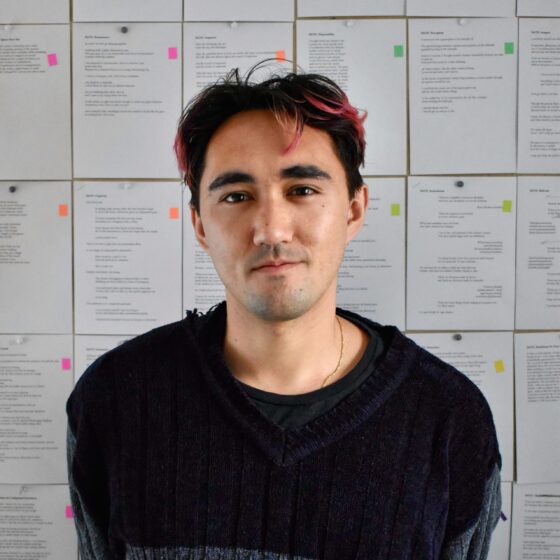 Rob Macaisa Colgate, a Filipino-American person, wears a dark long-sleeved top and stands in front of pages pinned on a wall. The tips of their short hair are red.