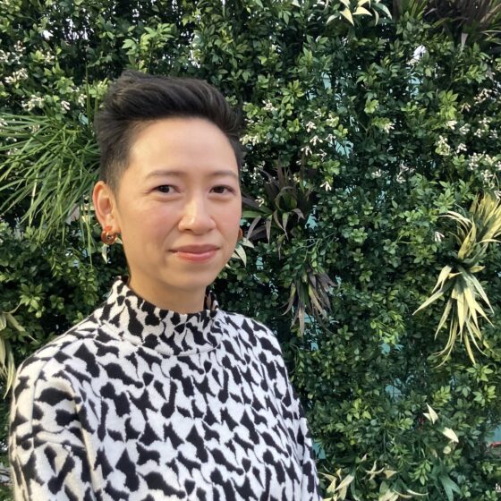 An Asian American woman with short-cut hair. She's wearing a white and black blouse and standing in front of a green bush
