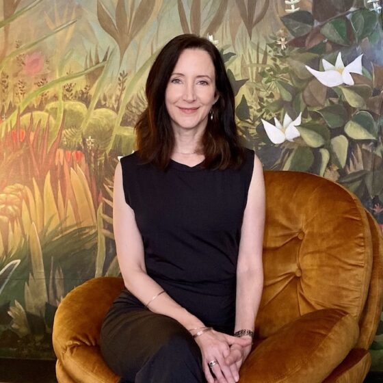 Author posing against a floral background with dark brown shoulder legth hair and all balck jumpsuit