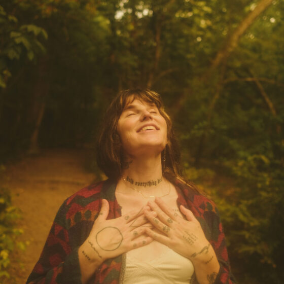 Rose Zinnia, a white woman with brown hair wearing a plaid shirt over a white tee, presses her hands over her heart and smiles at the sky. She's in a wooded area.