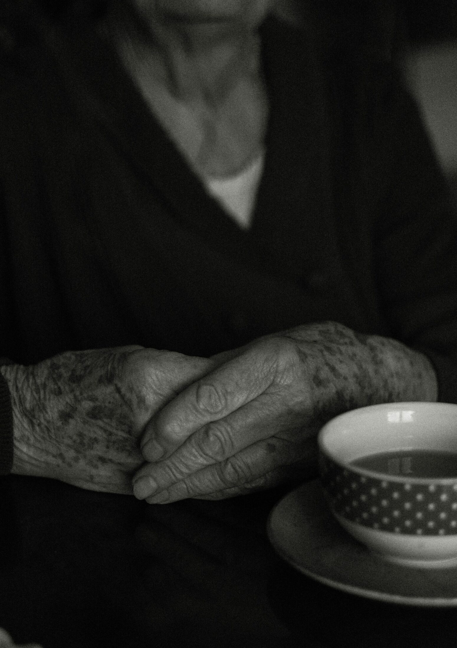 Black and white picture of an old woman with hands folded over one another in front of a teacup