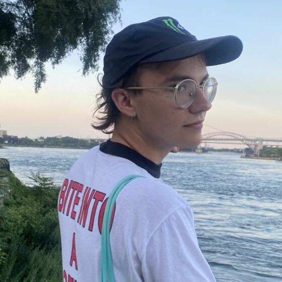 Theodore Heil, a white person with glasses and wearing a baseball cap, stands in front of a body of water and looks back at the camera.