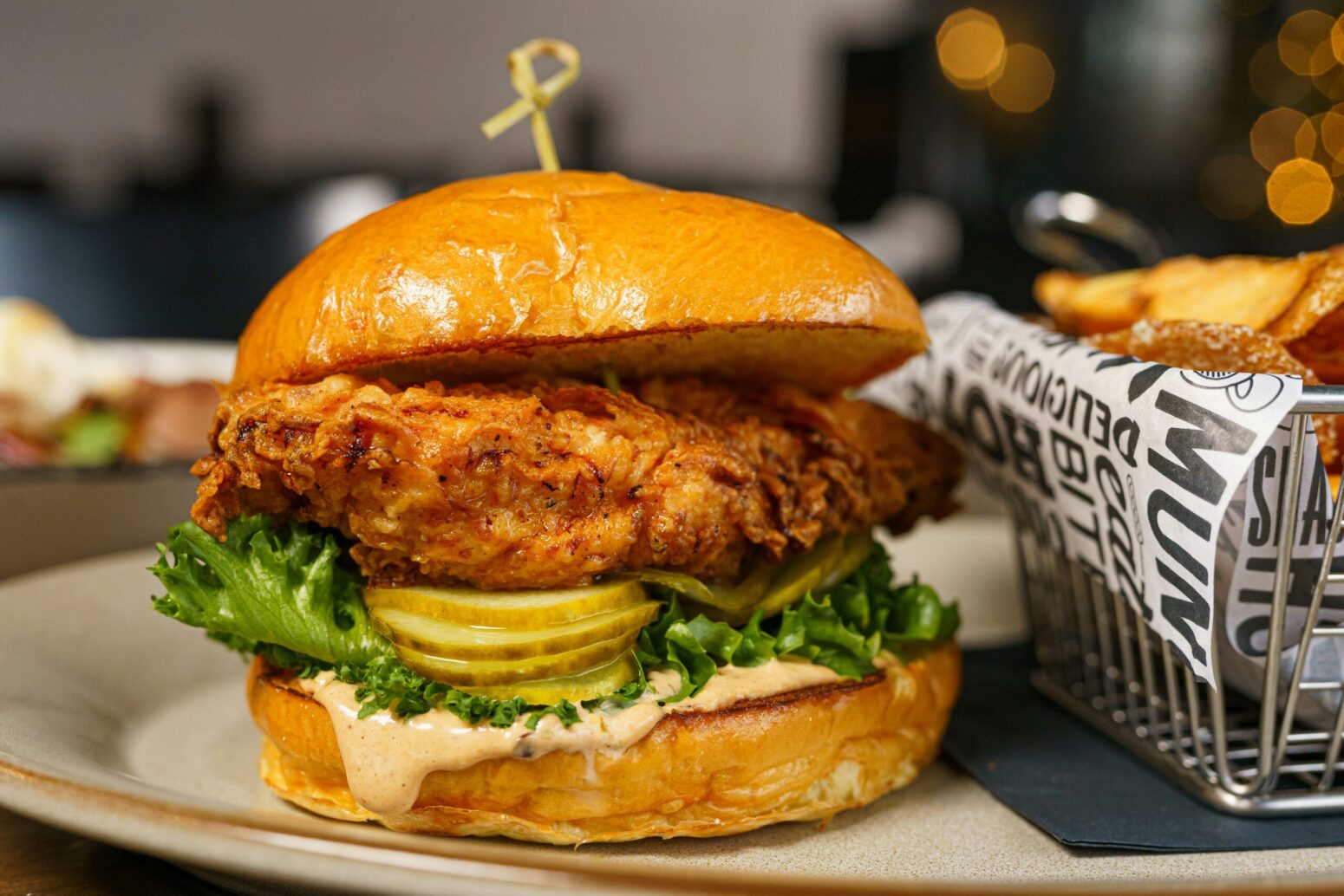 This photograph shows a chicken sandwich on a plate with a sauce, pickles, and lettuce on a brioche bun.