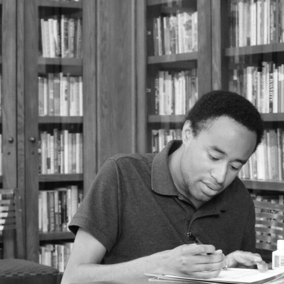 Prince Bush, a Black man with short hair, wears a polo and sits in front of some bookcases writing in a notebook.