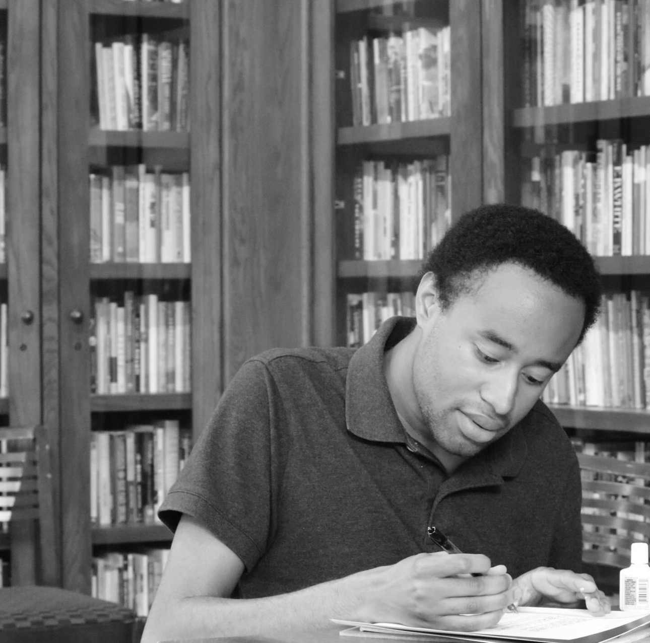 Prince Bush, a Black man with short hair, wears a polo and sits in front of some bookcases writing in a notebook.