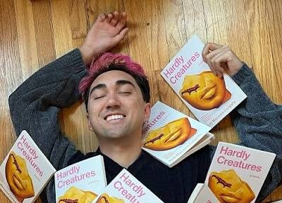 Rob Macaisa Colgate, a Filipino-American person with short hair with red tips, lies on a wooden floor, eyes closed, covered in copies of their book, "Hardly Creatures."