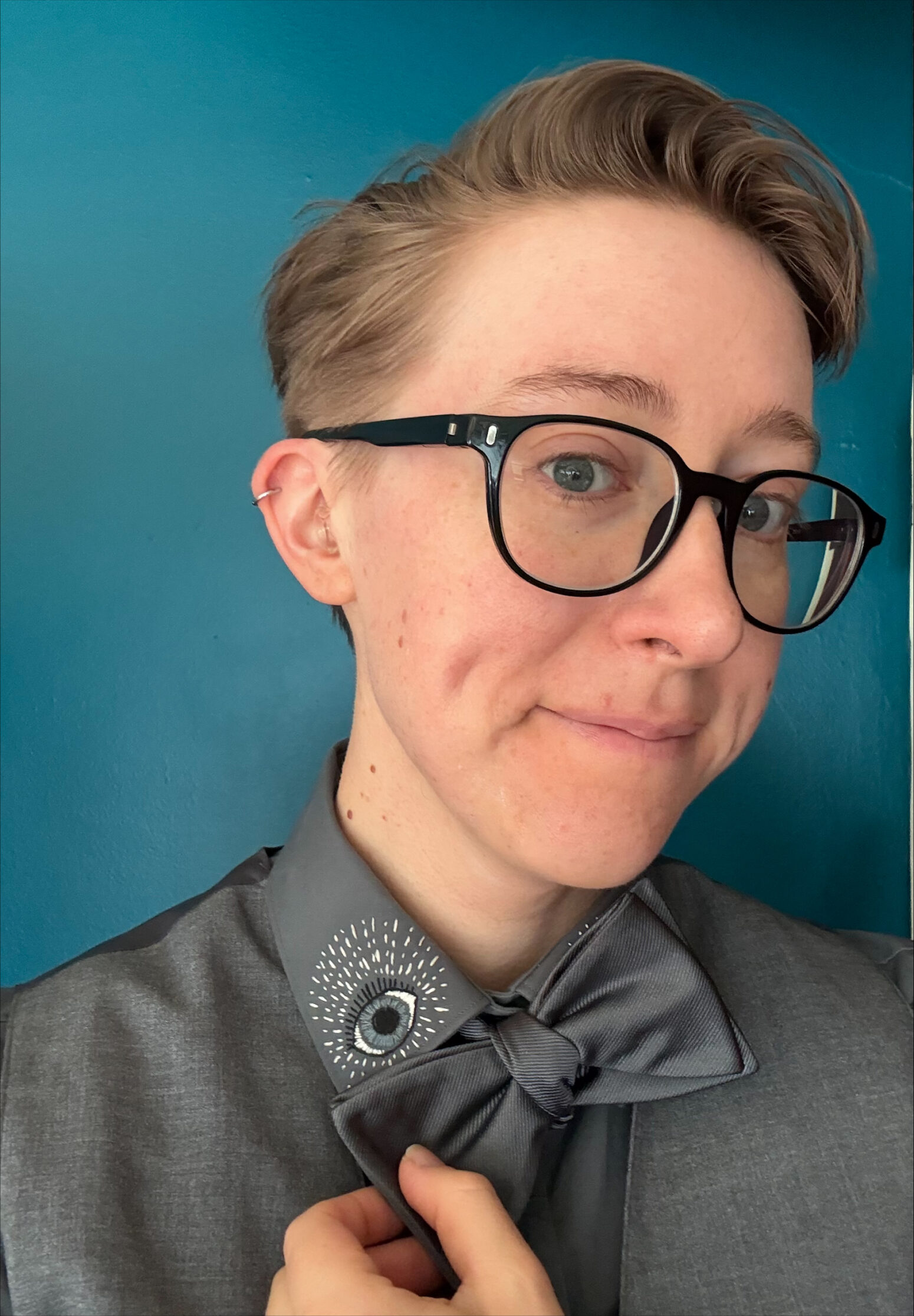 Lily Davenport, a white woman with short blond hair, wears black-rimmed glasses and a gray shirt and bow tie that she pulls down to reveal an eye embroidered on the collar of her shirt.