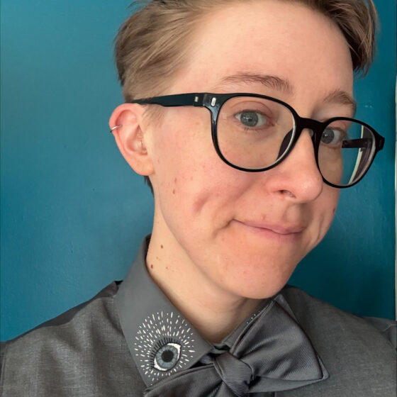 Lily Davenport, a white woman with short blond hair, wears black-rimmed glasses and a gray shirt and bow tie that she pulls down to reveal an eye embroidered on the collar of her shirt.
