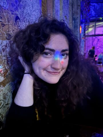 Anna Story, a young white woman with wavy brown hair, leans against a heavily graffitied wall and smiles at the camera. Rainbow lights are reflected on her face.
