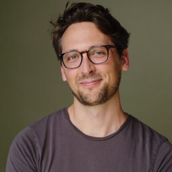 A light-skin man with short hair, black glasses, and a gray shirt poses with a slight grin in front of a solid-brown background.