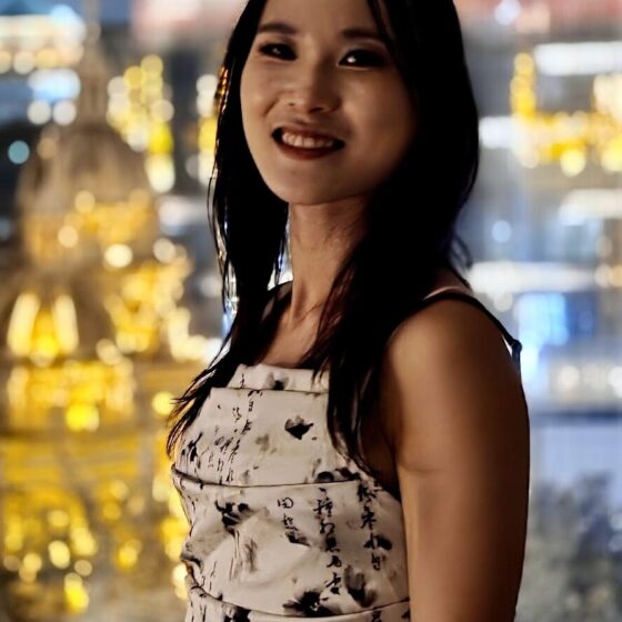 An Asian woman with shoulder length black hair stands in a white dress against a nighttime city skyline.