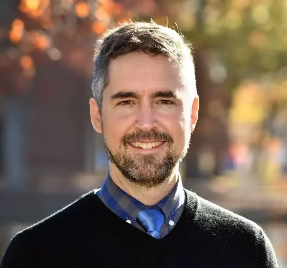 A man in a sweater and greying beard and hair smiling at the camera