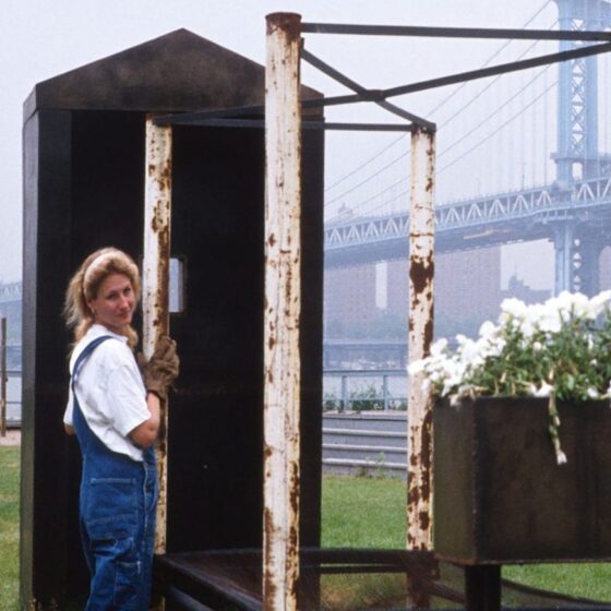 A blond woman with white shirt and overalls looking over her shoulder