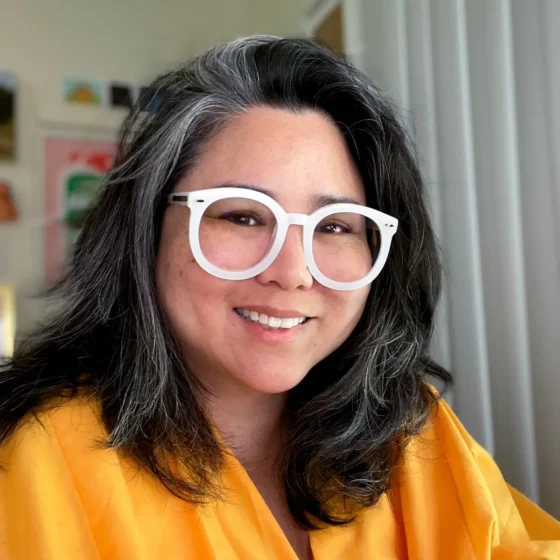 A woman with a gray streak and short black hair with white rimmed glasses wearing a yellow blouse smiling at the camera