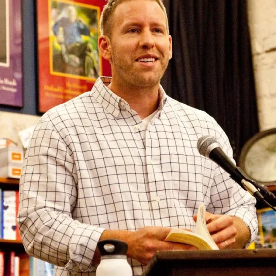 A blond man in a white checkered shirt in front of a mike stand
