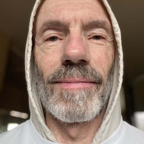 Peter Krumbach, a white man with a salt-and-pepper beard, wears a hood over his head and smiles.